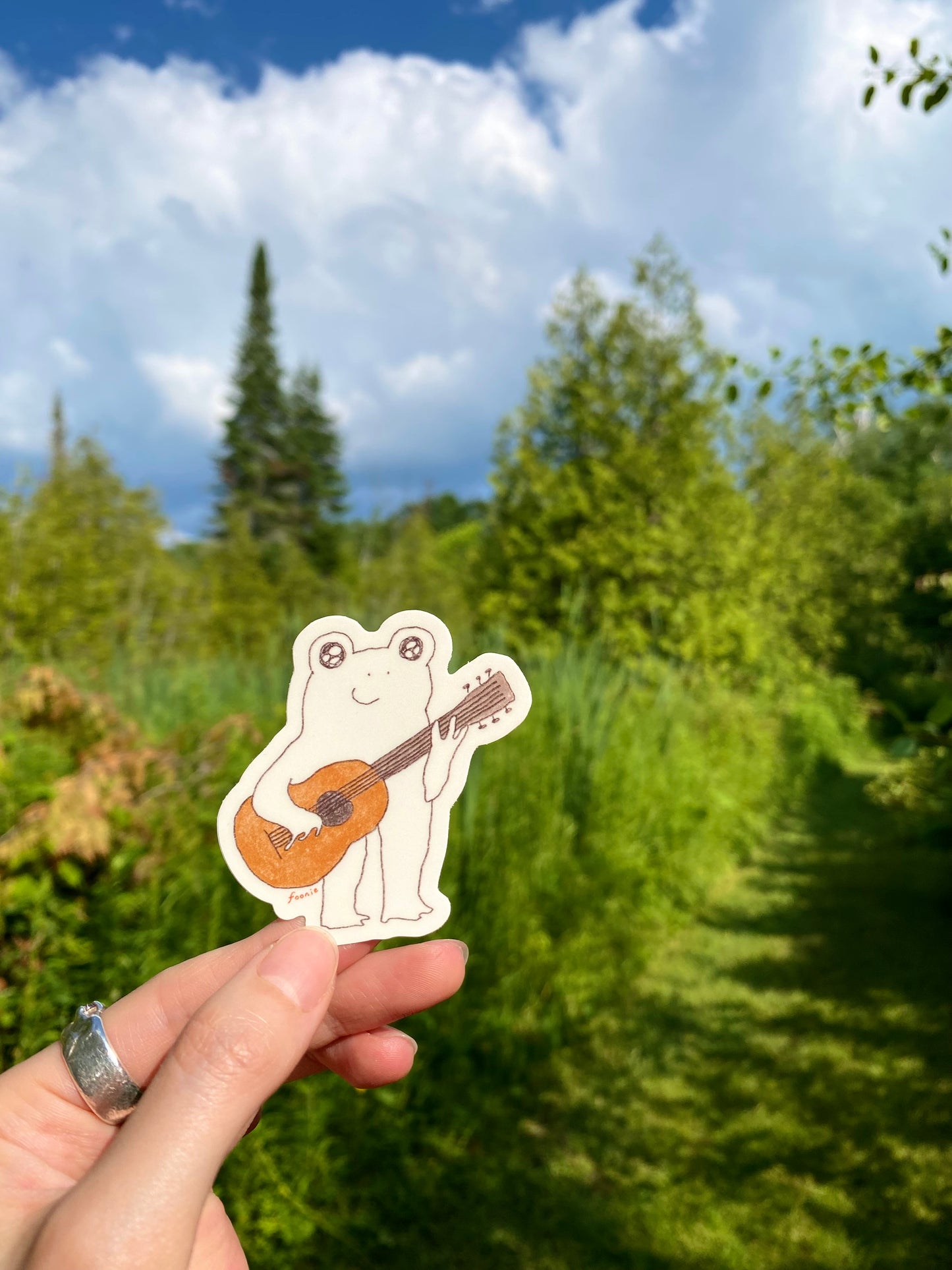 Hand holding a sticker of a frog playing a guitar against a natural background with trees and blue sky.