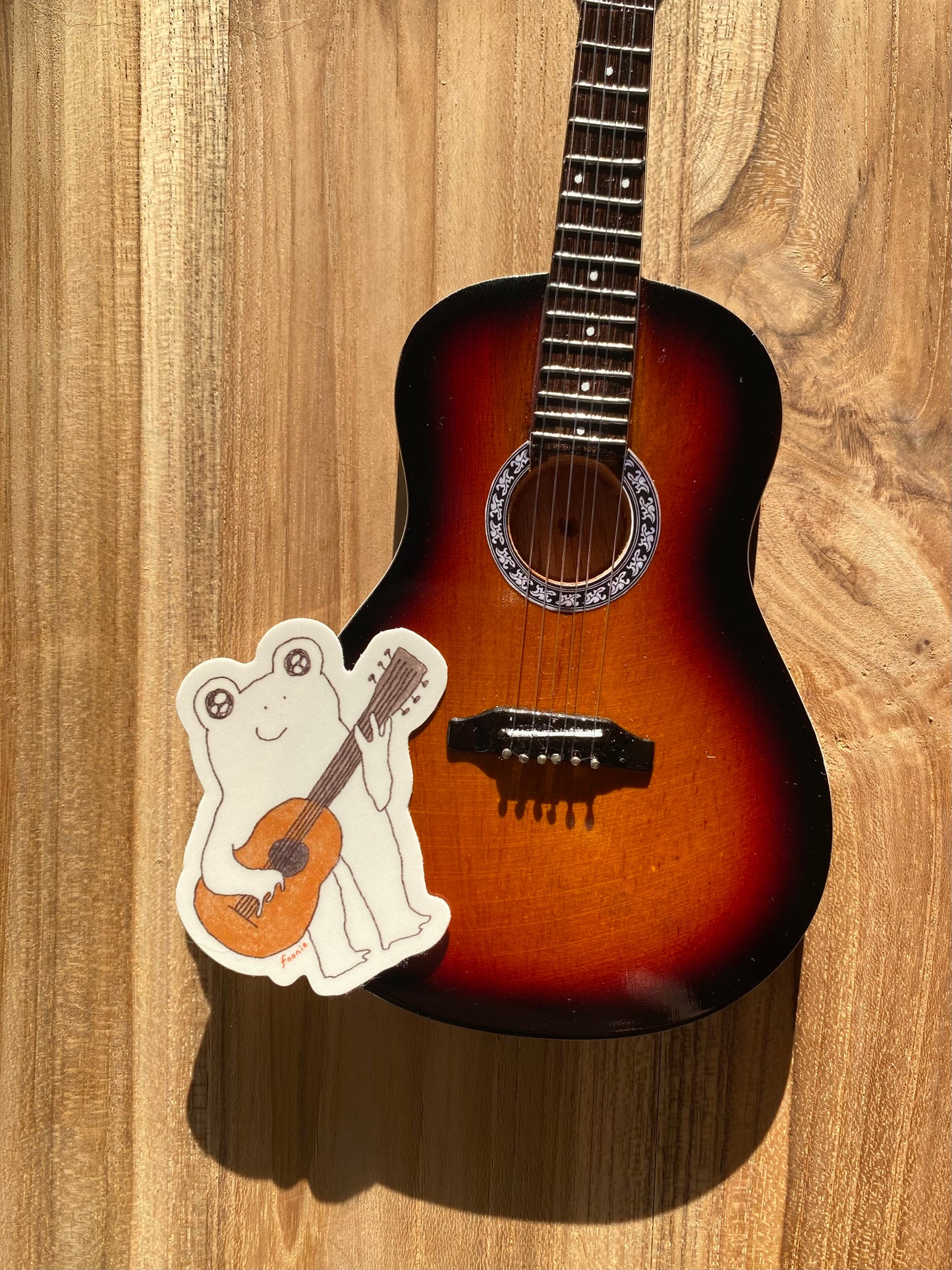 Mini acoustic guitar on a wooden surface with a sticker of a frog playing a guitar.