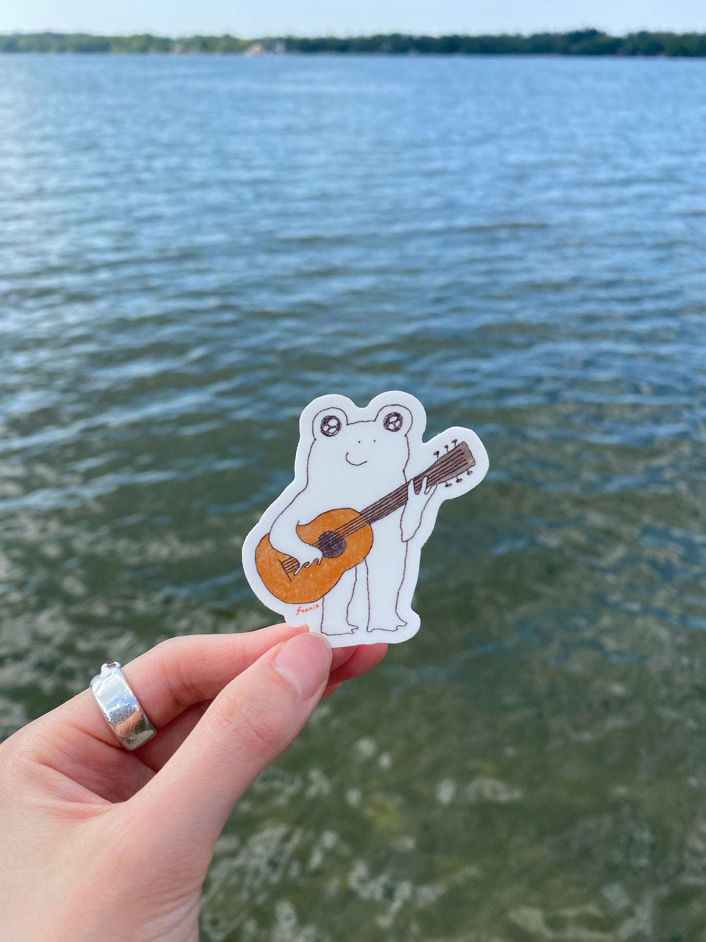 Hand holding a sticker of a frog playing a guitar against a water background