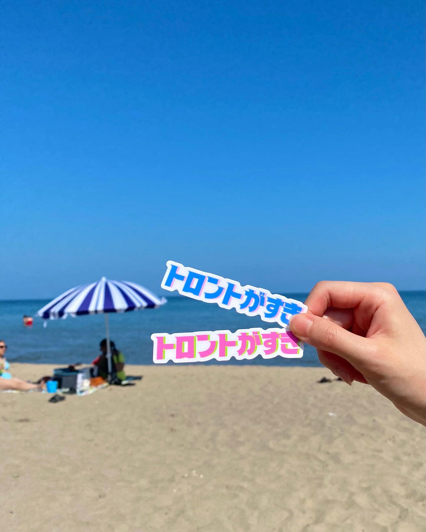 Beach scene with hand holding two stickers that say "I love Toronto" in Japanese in front of a clear blue sky.