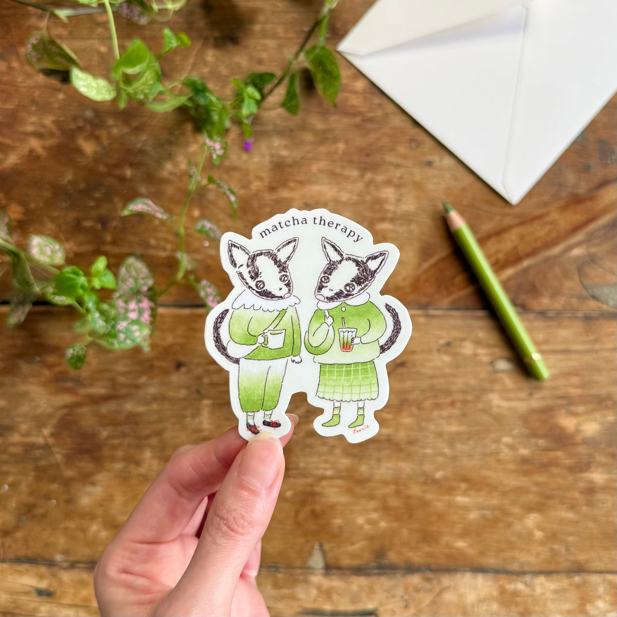 A hand holds a die-cut sticker of two illustrated creatures dressed in green outfits holding matcha cups, with the text 'matcha therapy' above them. The scene is set on a rustic wooden table with a green pencil, a white envelope, and green leaves in the background.