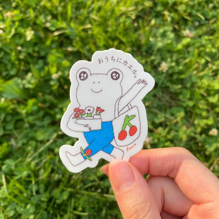 a sticker with the illustration of a cute frog with flowers and a cherry tote bag held by a hand