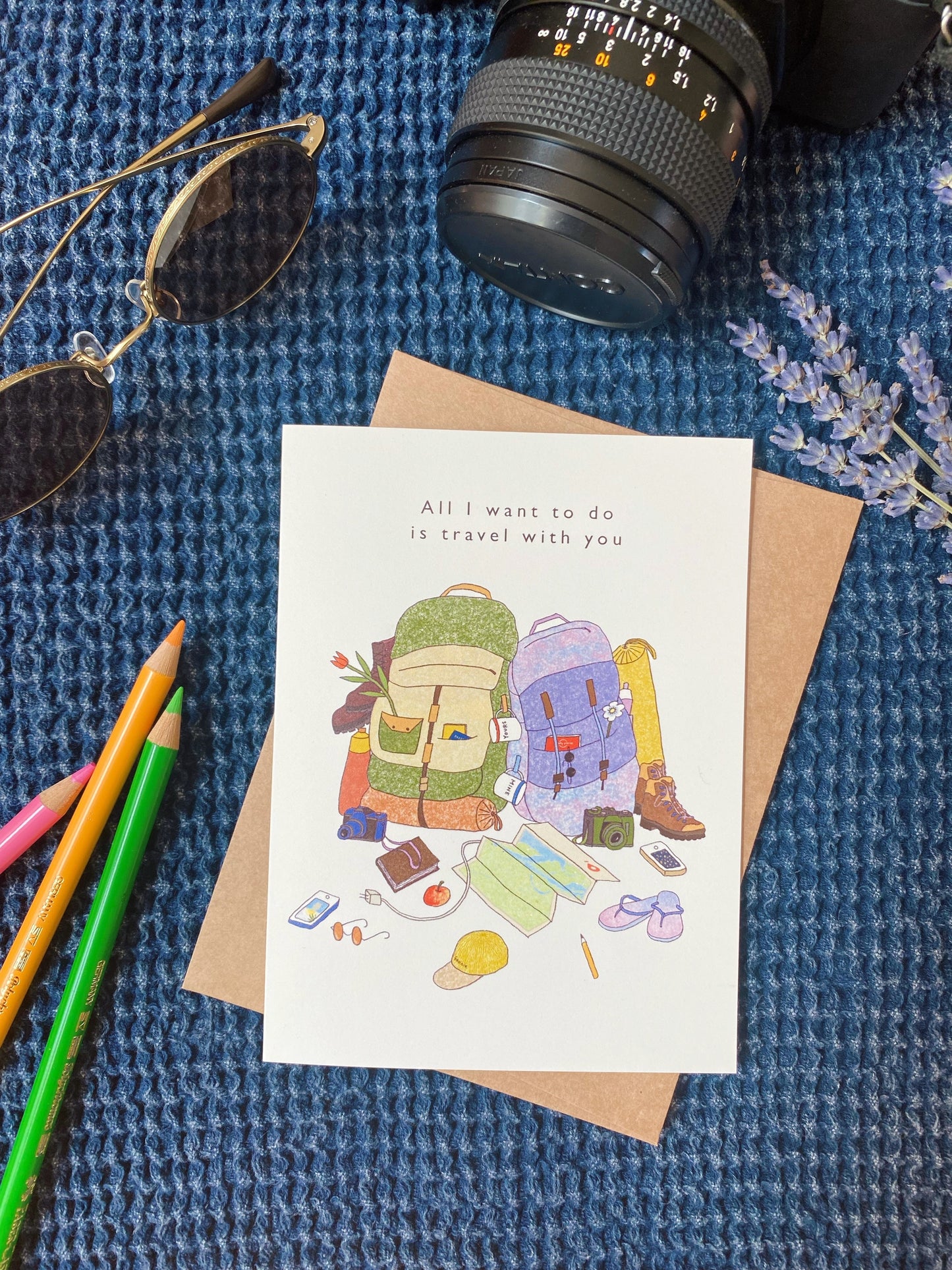 Just because Card that says “All I want to do is travel with you” with the illustration of two backpacks and travelling goods such as maps, sunglasses, books, yoga mats, phones, and beach sandals.