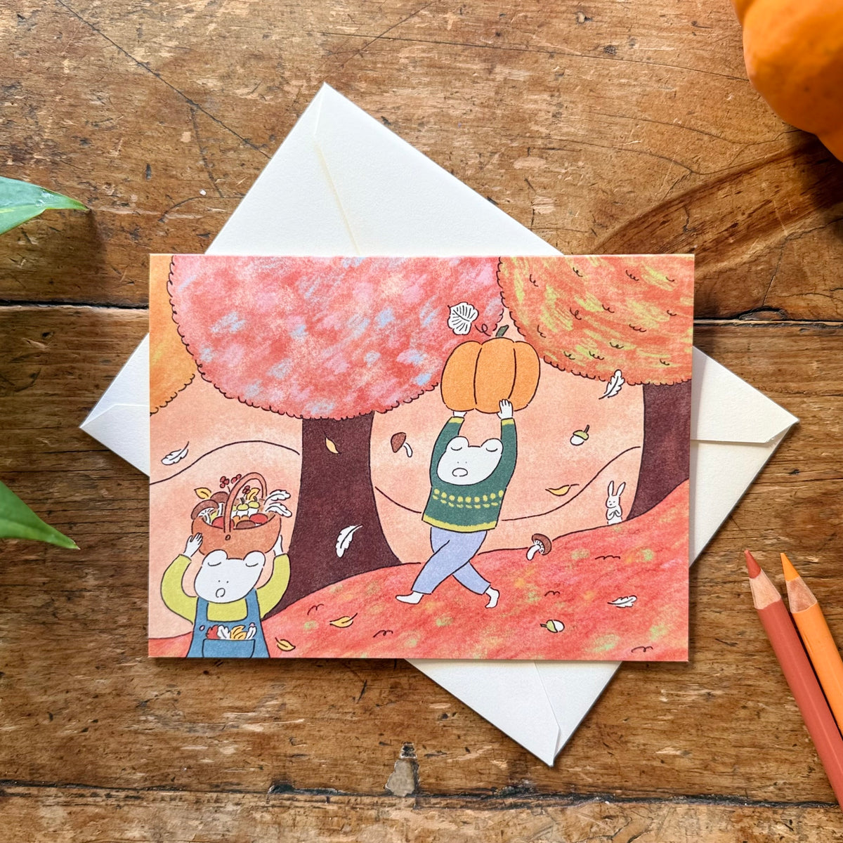 An autumn themed greeting card with a cream white envelope, placed on a rustic wooden table together. The card depicts a hand-drawn autumn scene with two frogs carrying a pumpkin and baskets full of acorns and mushrooms.  