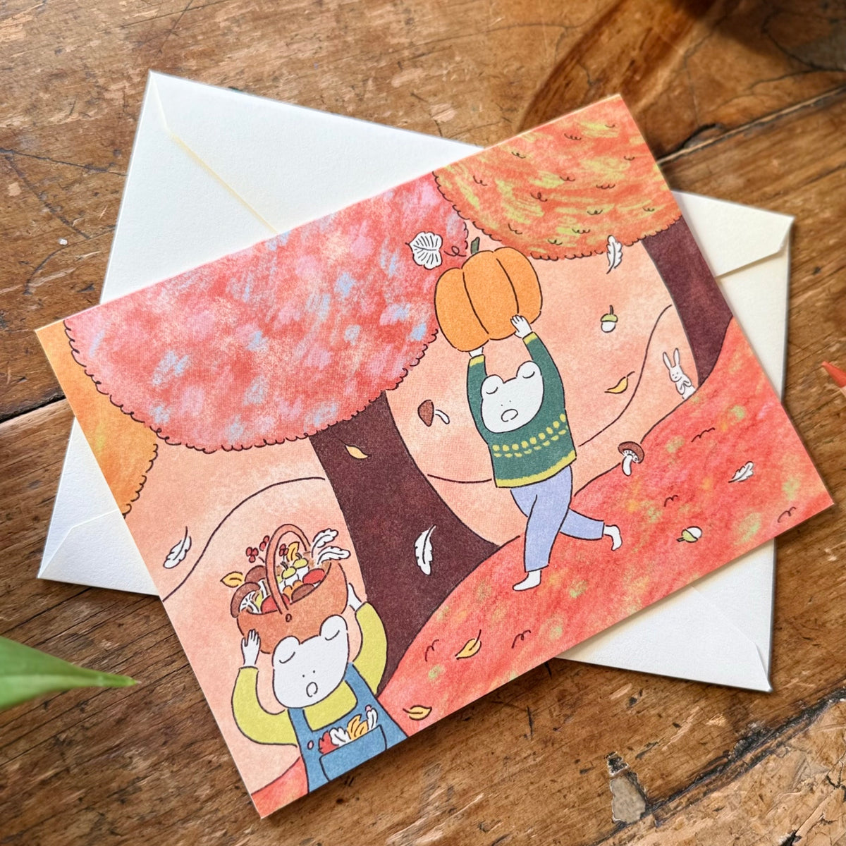 A fall themed greeting card with a frog carrying a pumpkin and the other frog holding a basket of fall produce. They are surrounded by autumn trees. 