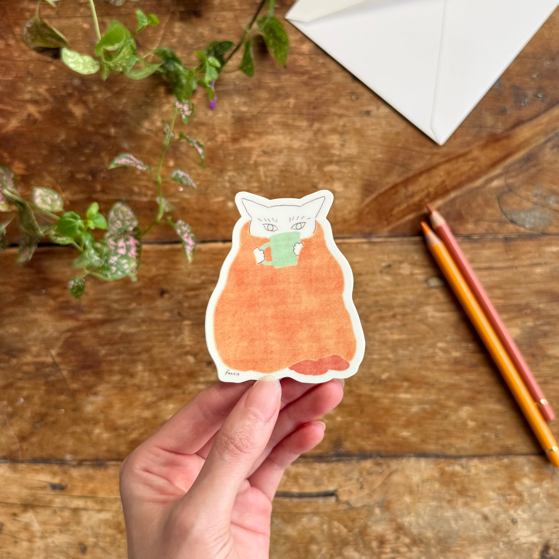 A hand holds a die-cut sticker of a white cat with a green mug, wrapped in a blanket, shown on a rustic wooden table with plants and colored pencils.
