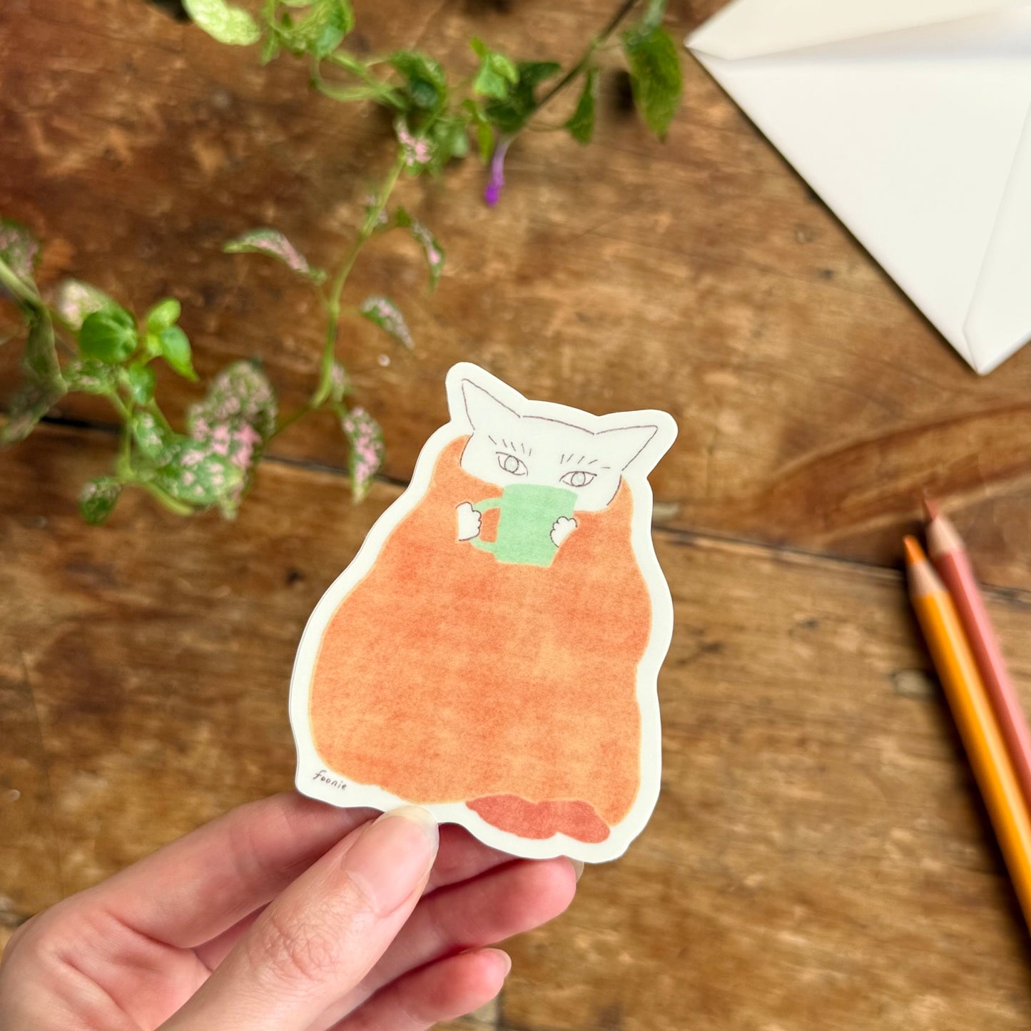 A hand holds a vinyl sticker with the illustration of a cute cat wrapped in a orange blanket, set against a rustic wooden tabletop with plants and colored pencils in the background.