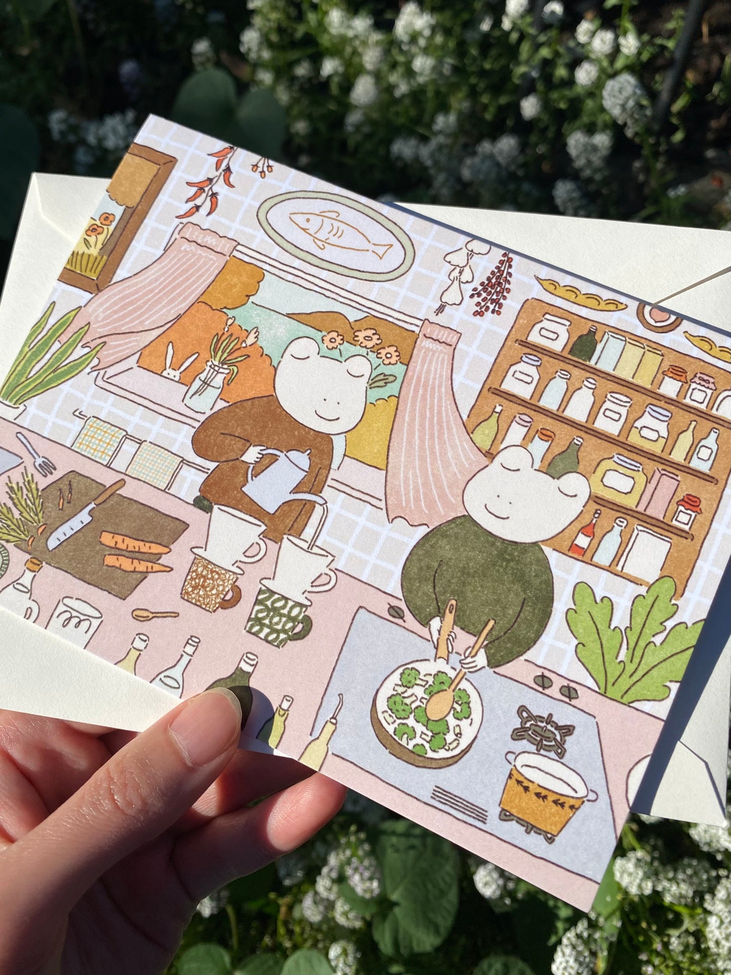 Illustrated greeting card with two frog characters in a kitchen setting, held by a hand with a blurred green background.