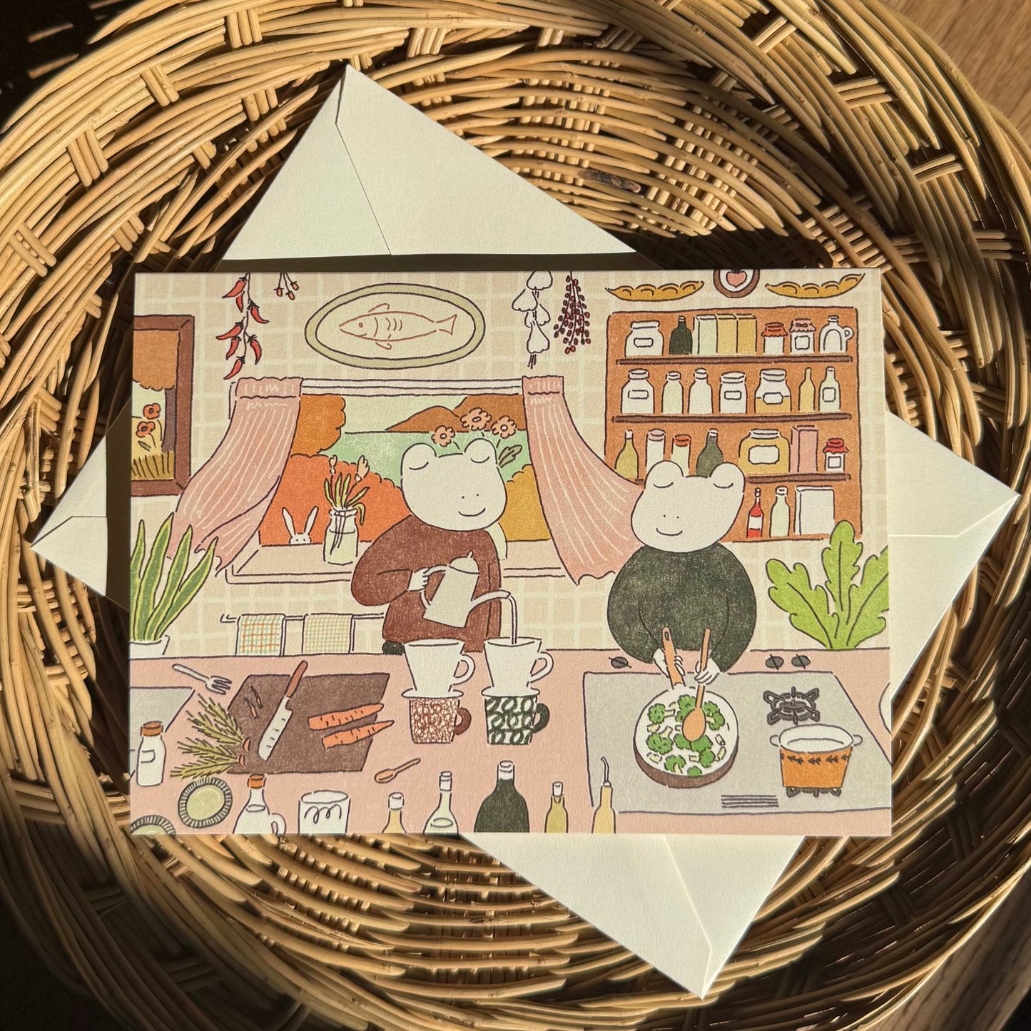 Illustrated card with two frog characters in a kitchen setting, placed on a woven basket.
