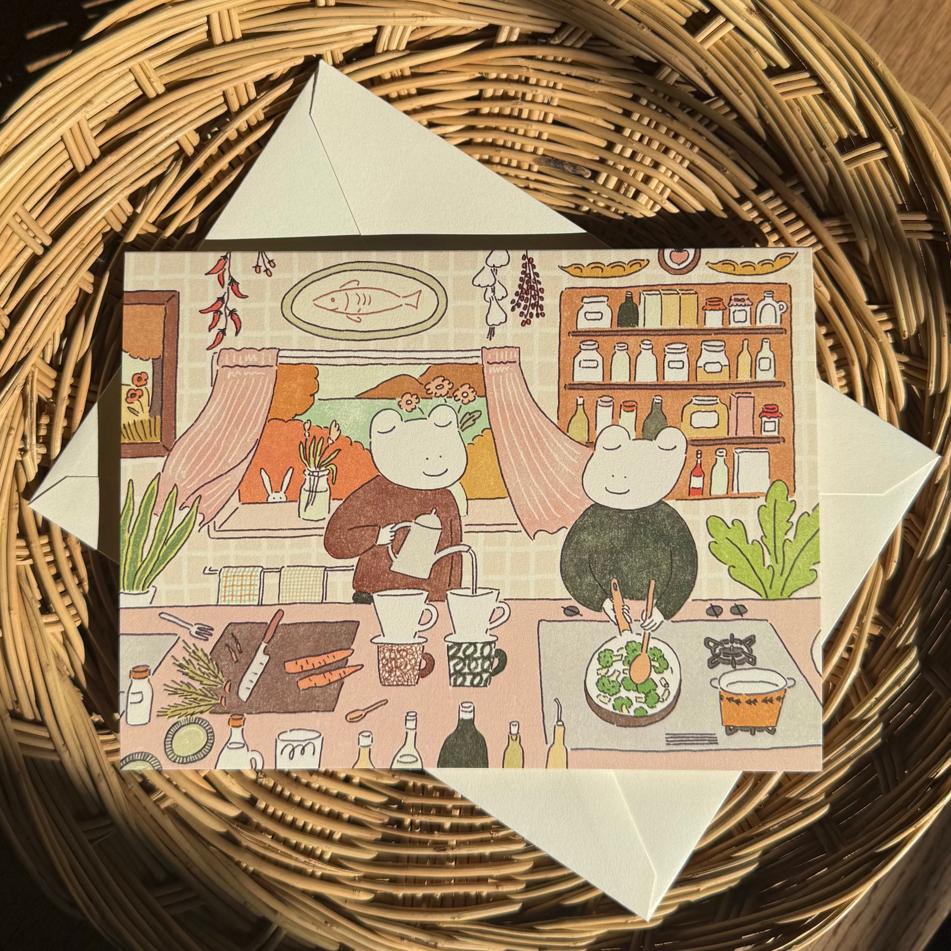 Illustrated card with two frog characters in a kitchen setting, placed on a woven basket.