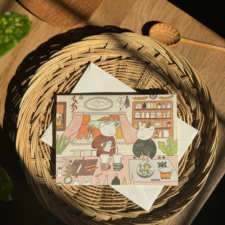 Illustrated card with two frogs in a kitchen scene on a woven basket