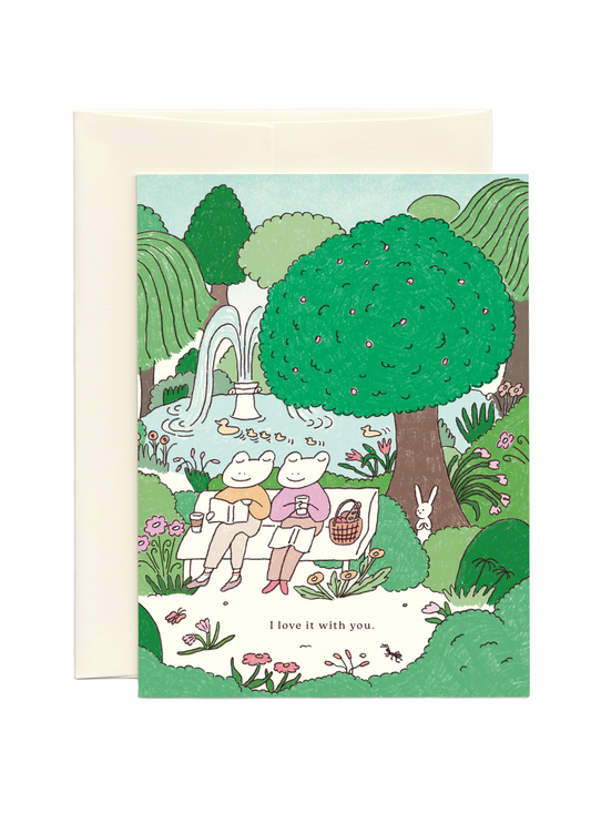 Greeting card with illustration of two frogs sitting on a bench in a park reading and text 'I love it with you.'