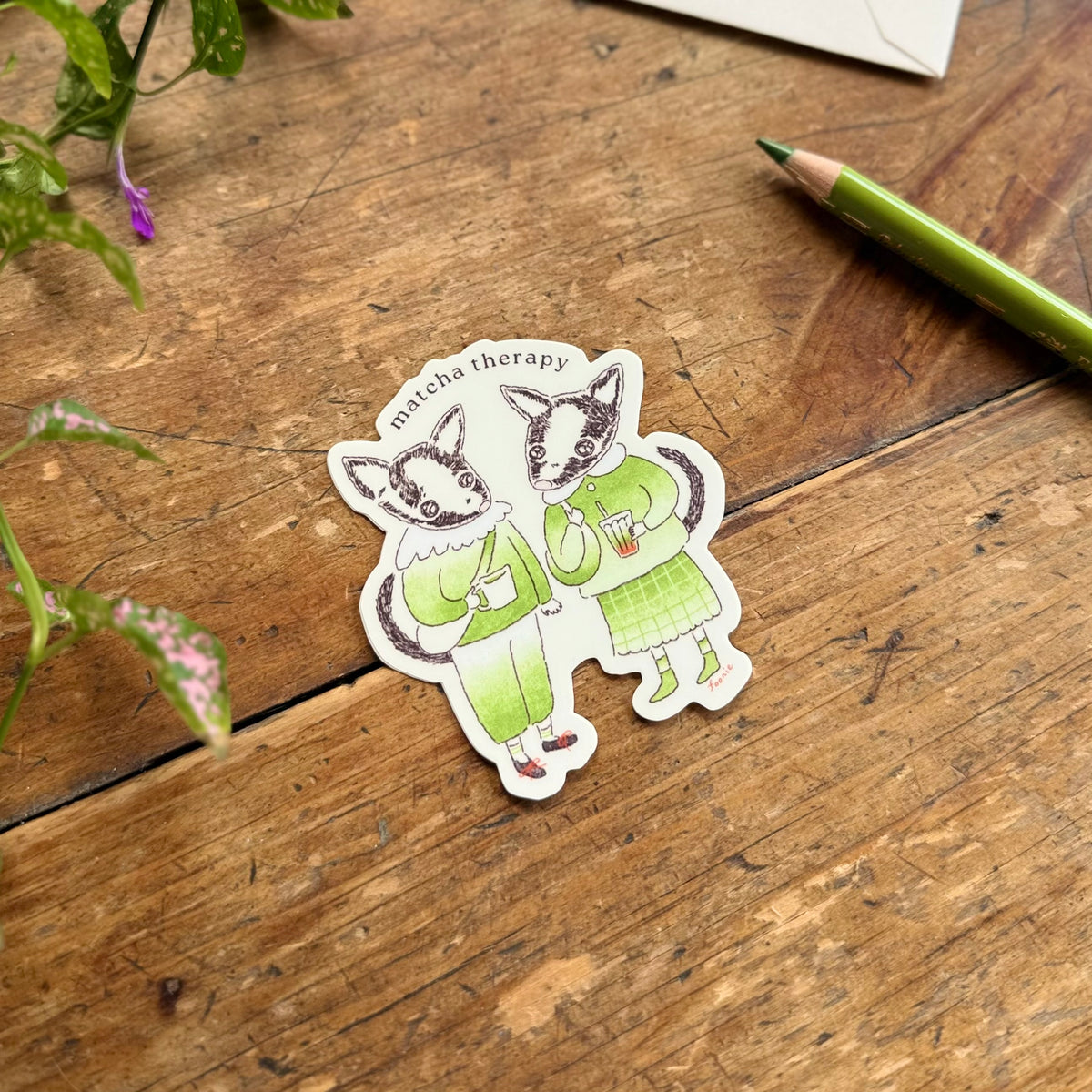 A vinyl sticker of two civets dressed in green outfits, placed on a rustic wooden tabletop with a green pencil and plant leaves nearby. The sticker reads 'matcha therapy'.