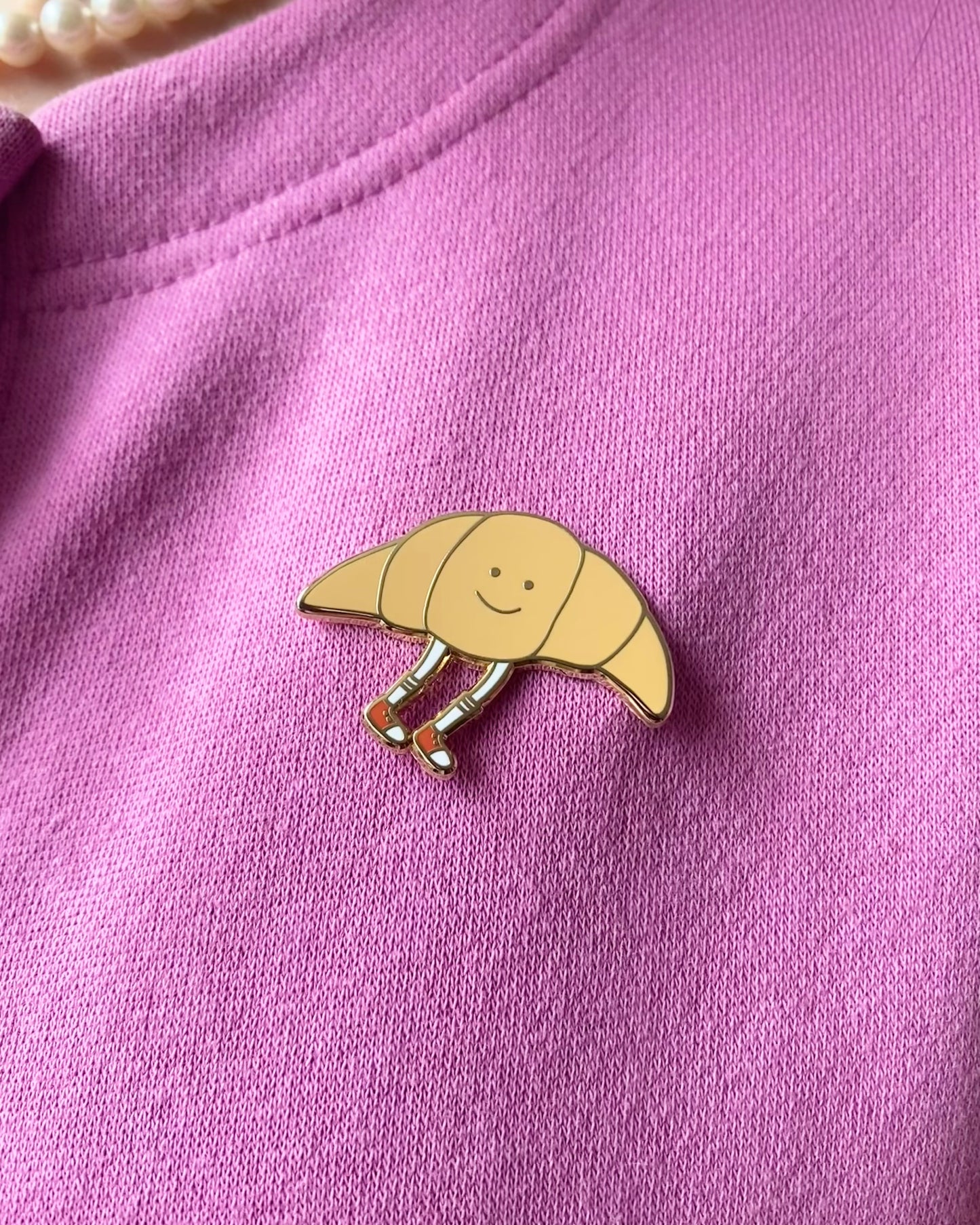 A cute croissant enamel pin on a pink sweater