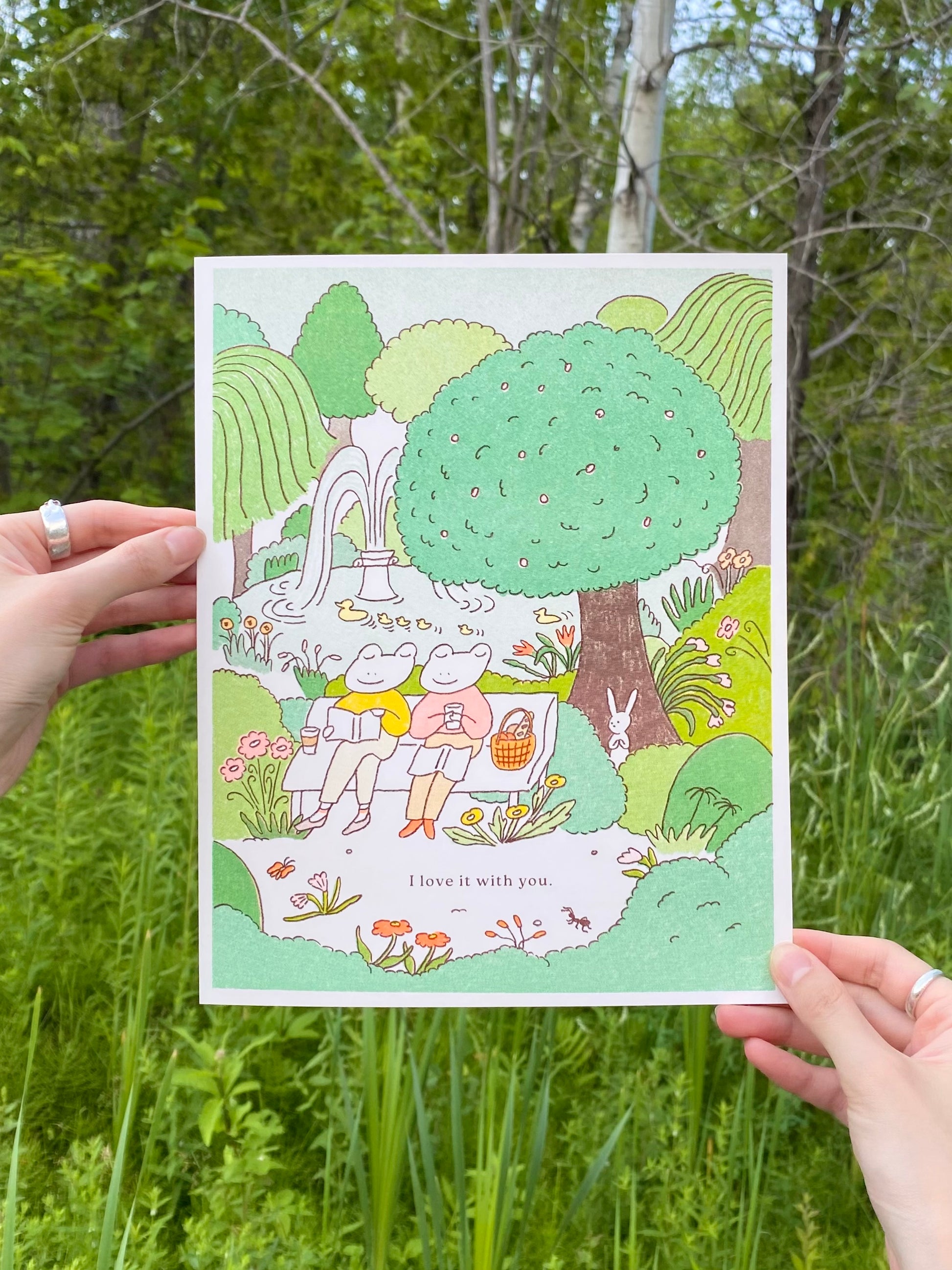 two hands holding a cute art print with the illustration of two frogs sitting on a bench 