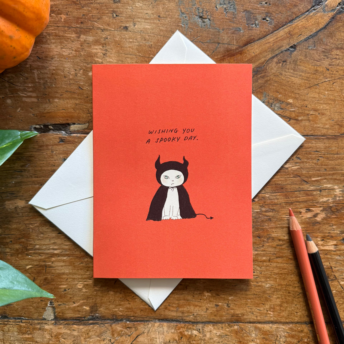 An orange greeting card with the illustration of a cat wearing a black devil halloween costume, with the text 'WISHING YOU A SPOOKY DAY.' 