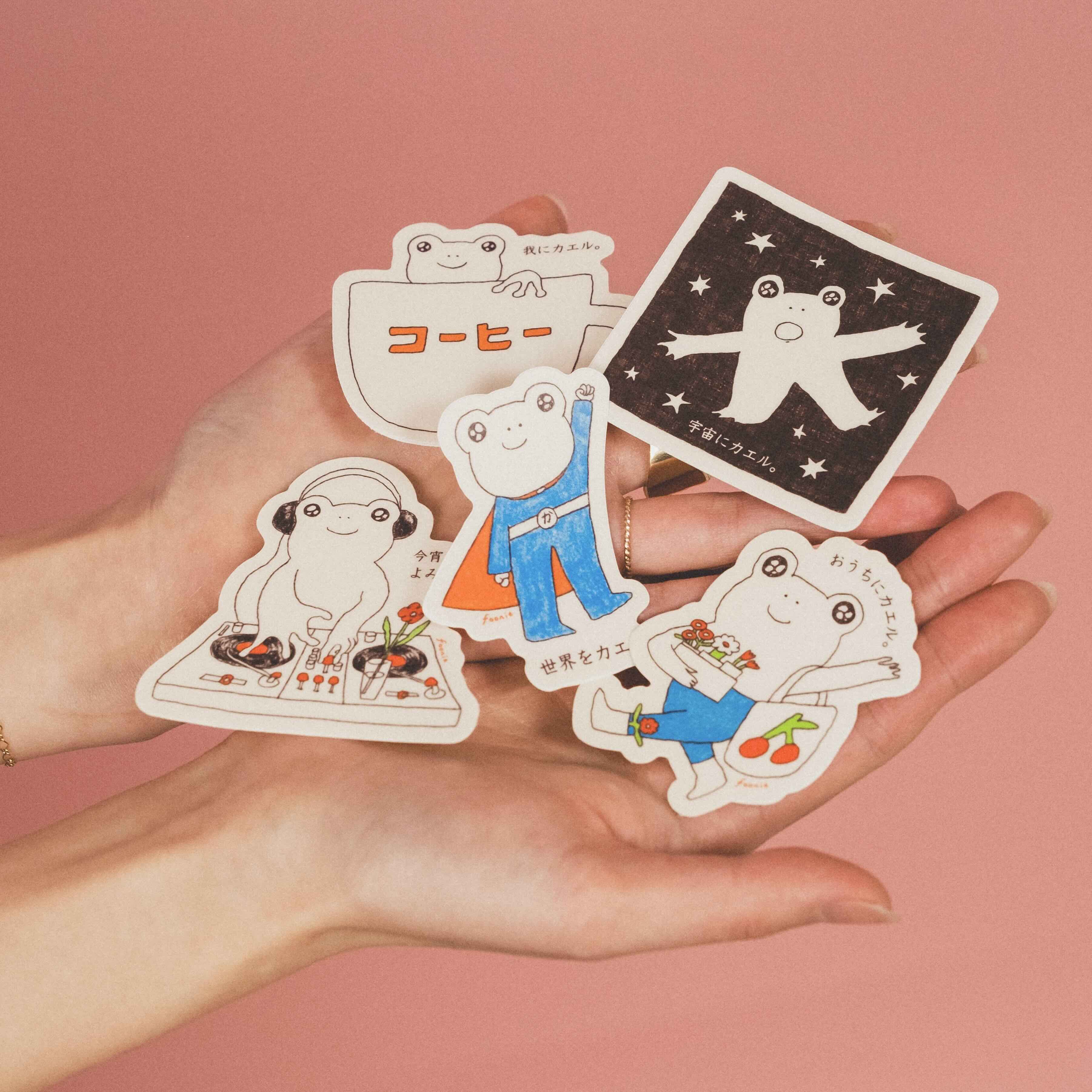 Hands holding five vinyl cartoon frog stickers against a pink background