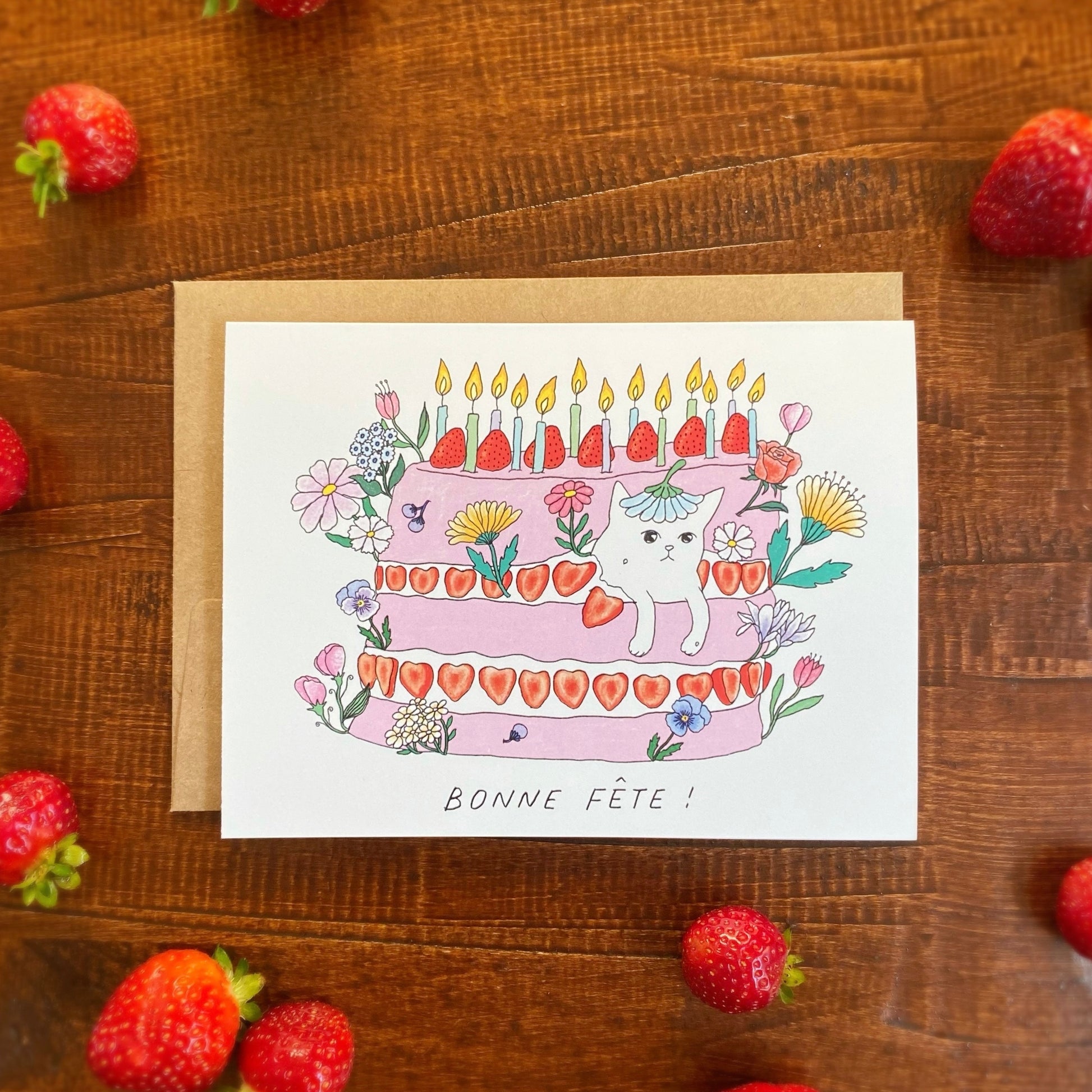 Cute birthday card of a white cat coming out of a pink birthday cake with lots of strawberries and flowers