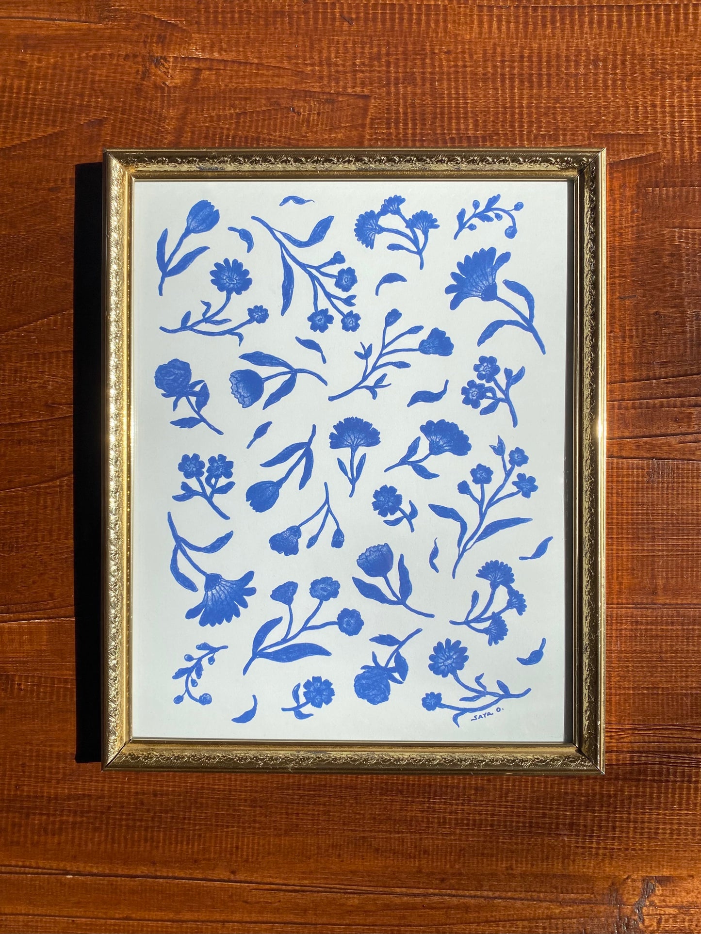 A classic porcelain-style floral print with blue flowers on a natural white background in a gold frame