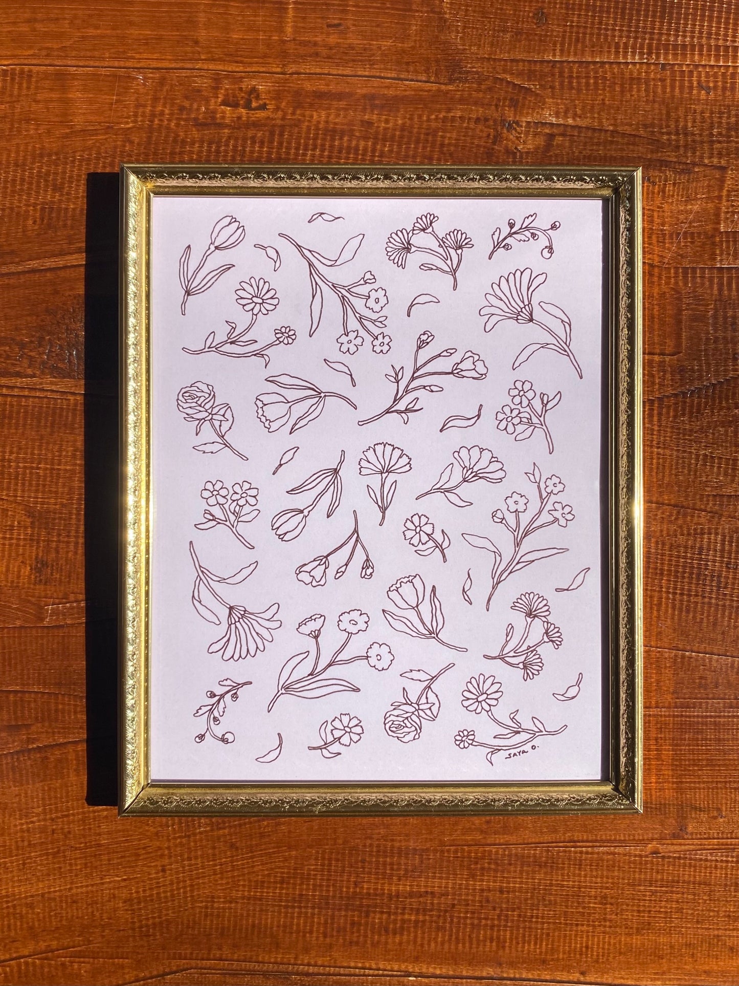 A floral textile print of the simple illustration of various flowers on a soft pink background in a gold frame